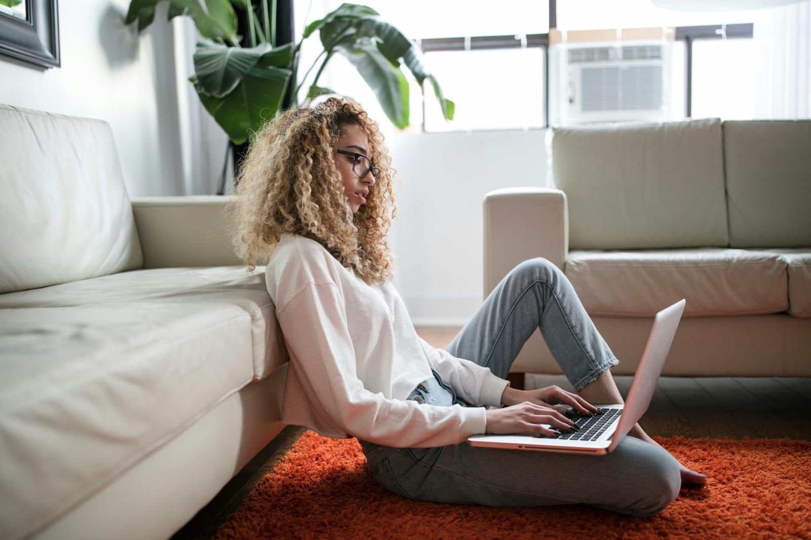 A woman sits on the floor of her living room and works on a laptop